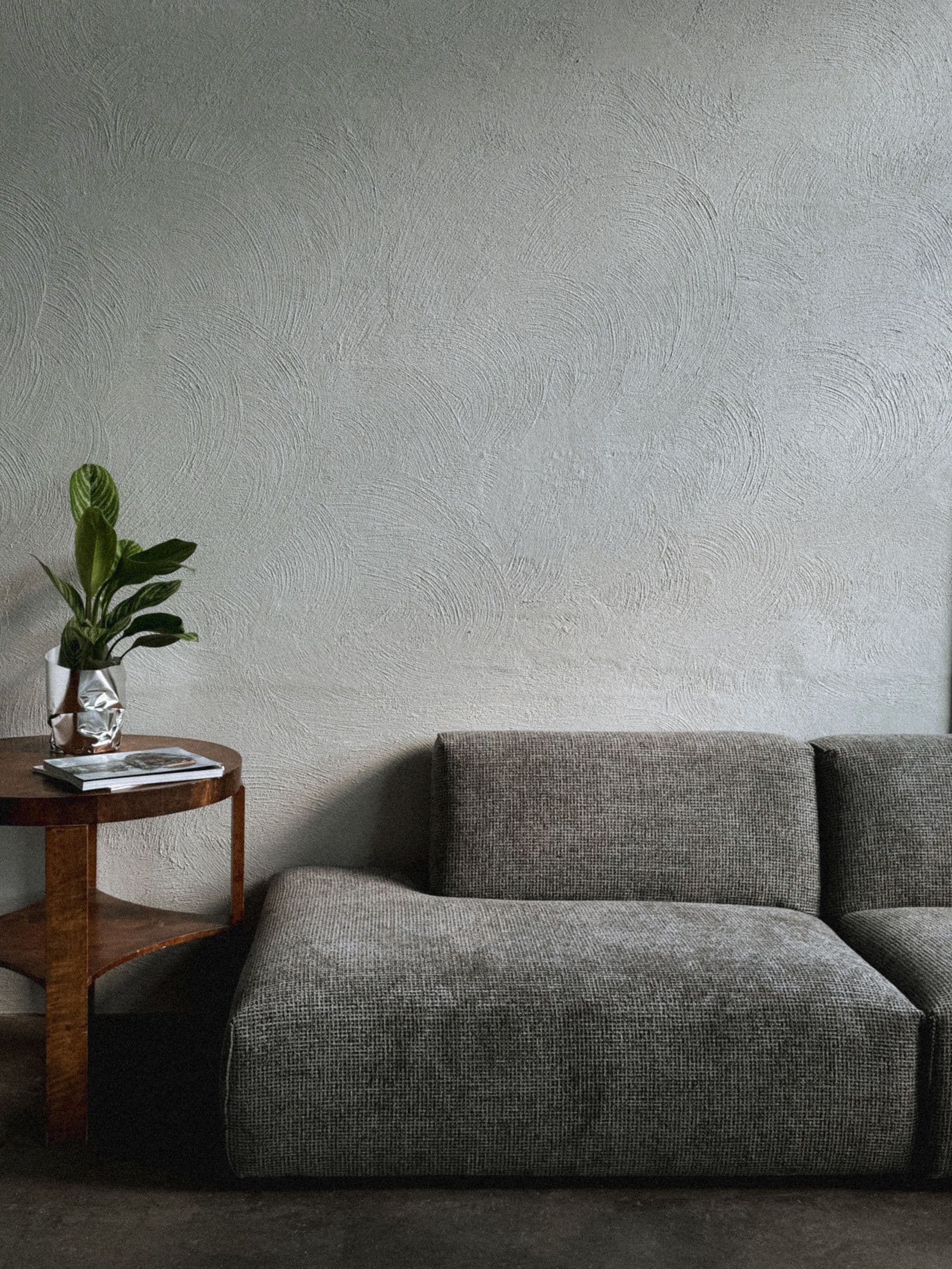 A calm living room — a low grey sofa and a wood side table against a hand-textured sage plaster wall.
